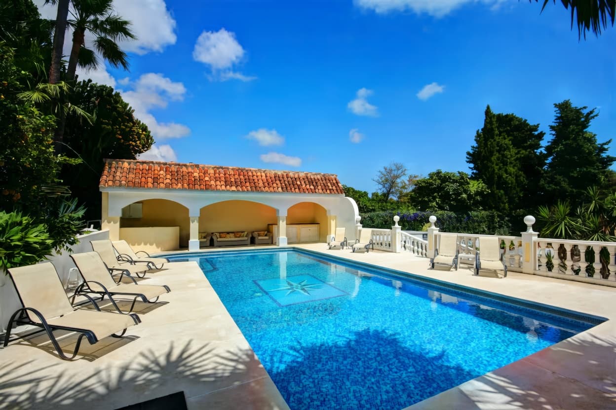 Piscina azul con tumbonas, cabaña de tejas rojas y vegetación bajo un cielo azul despejado.