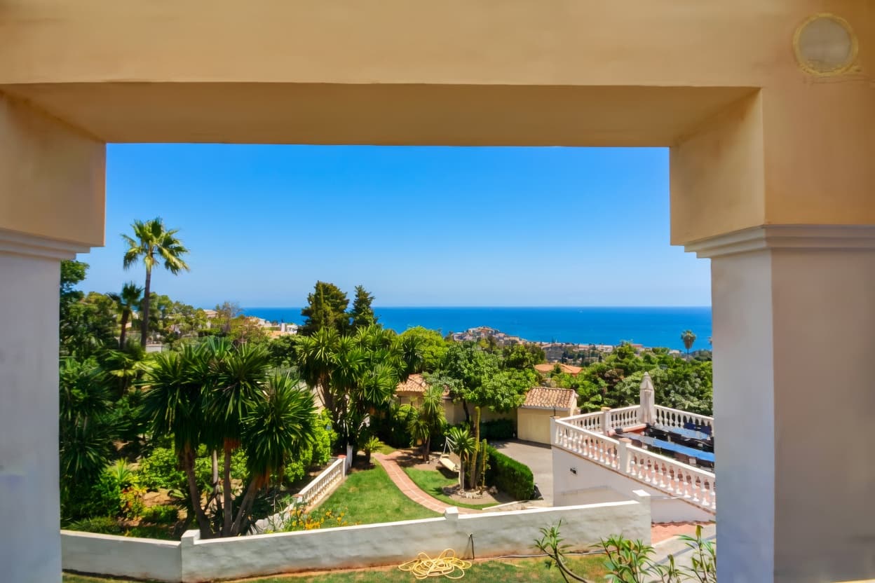 Vista enmarcada de un jardín con palmeras y terraza blanca frente al mar azul.